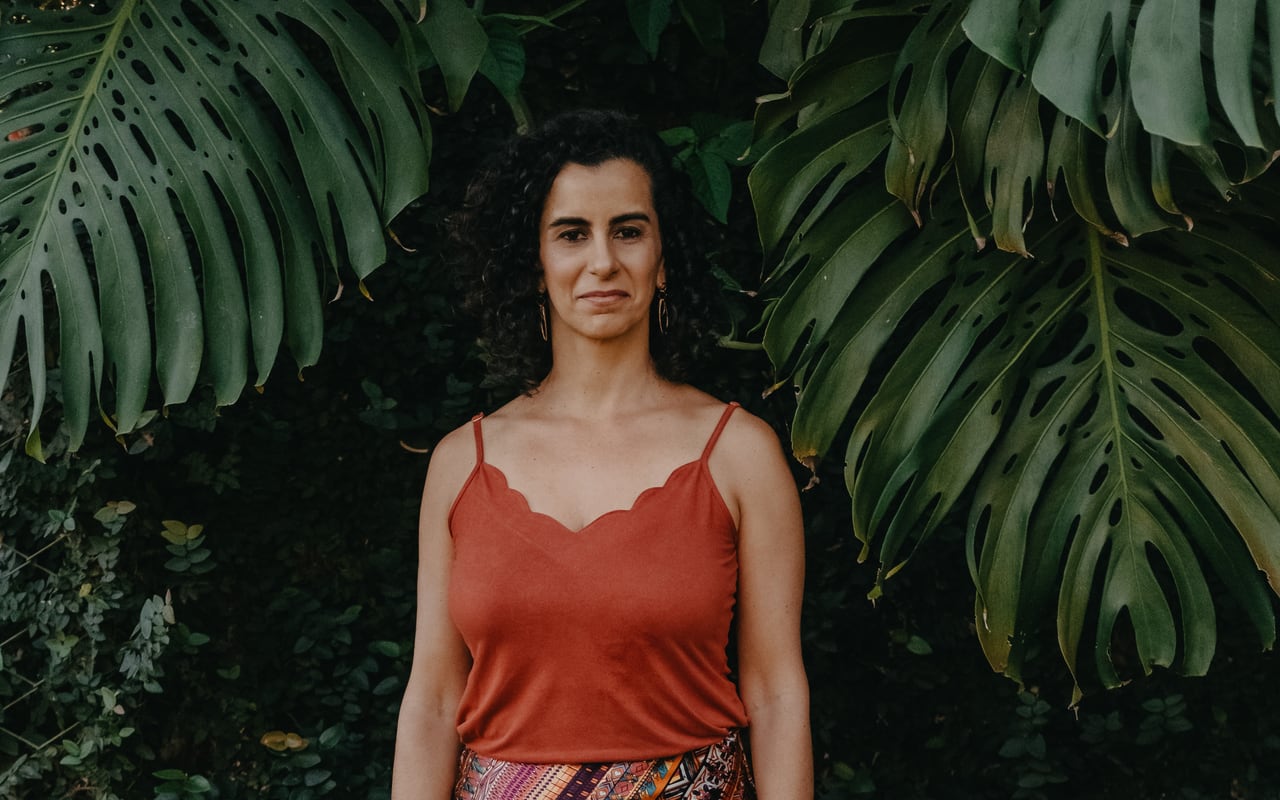 Lu Cerqueira em pé, serena e presente, entre folhas verdes